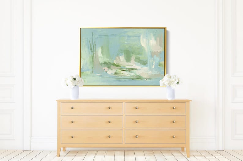 May include: An abstract painting in a gold frame hangs above a light wood dresser. The painting features soft blues, greens, and white brushstrokes. Two white vases with white flowers sit on the dresser, adding a touch of elegance.