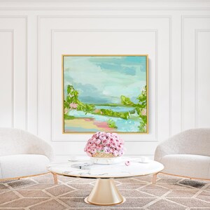 May include: Two white armchairs with gold trim face each other across a round marble coffee table with a gold base. A vase filled with pink flowers sits on the table. A painting of a landscape with a blue sky, green trees, and a river is hanging on the wall behind the chairs.
