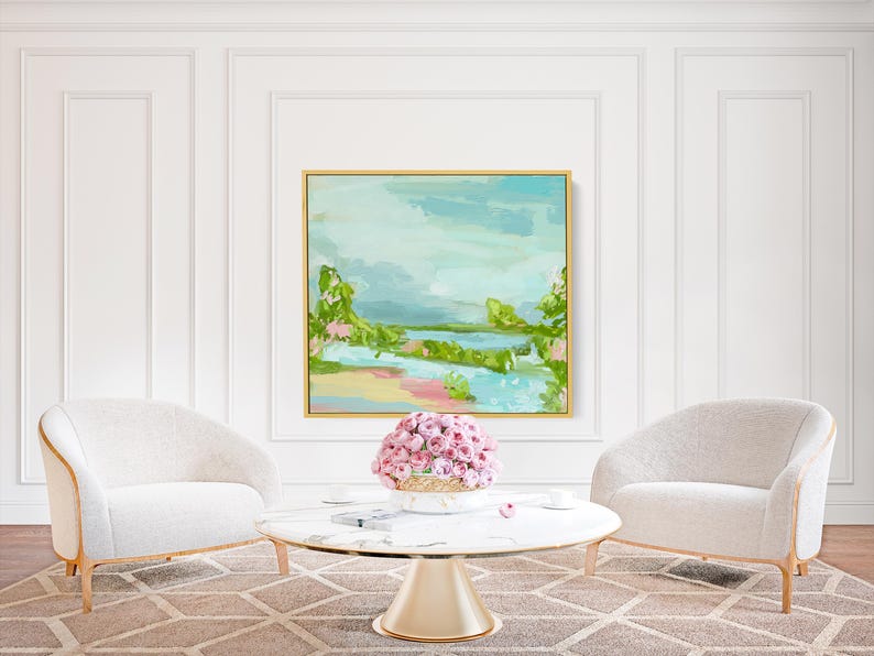 May include: Two white armchairs with gold trim face each other across a round marble coffee table with a gold base. A vase filled with pink flowers sits on the table. A painting of a landscape with a blue sky, green trees, and a river is hanging on the wall behind the chairs.