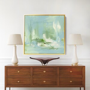 May include: An abstract painting in a gold frame, featuring shades of blue, green, and white. The artwork hangs above a dark wood dresser with six drawers and gold hardware. Two white lamps with beige shades flank the painting.