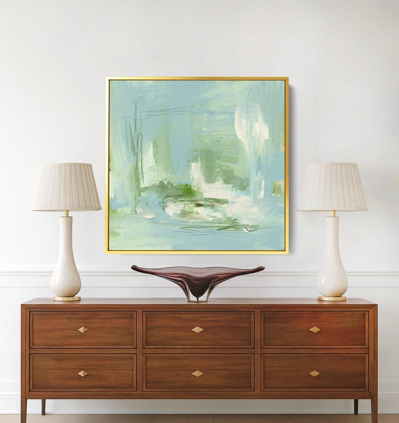 May include: An abstract painting in a gold frame, featuring shades of blue, green, and white. The artwork hangs above a dark wood dresser with six drawers and gold hardware. Two white lamps with beige shades flank the painting.