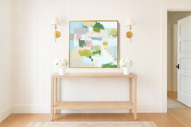May include: An abstract painting in a gold frame hangs above a light wood console table. The artwork features green, pink, and blue brushstrokes. Two white vases with flowers sit on the table, and wall sconces flank the artwork.