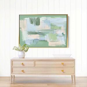May include: Abstract painting in a gold frame, with green, blue, and beige brushstrokes. It hangs above a light wood dresser with gold knobs. A white vase with flowers is on the dresser, creating a serene scene.
