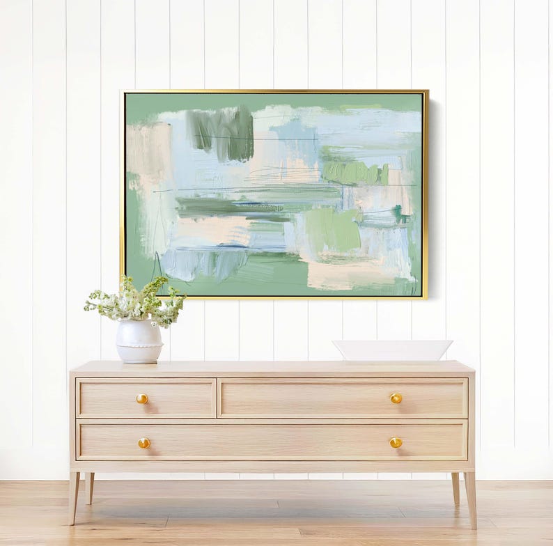 May include: Abstract painting in a gold frame, with green, blue, and beige brushstrokes. It hangs above a light wood dresser with gold knobs. A white vase with flowers is on the dresser, creating a serene scene.