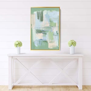 May include: Abstract art print in a gold frame. The artwork features brushstrokes in shades of green, blue, and beige on a green background. The painting is displayed above a white console table with a white vase and green flowers on each side.