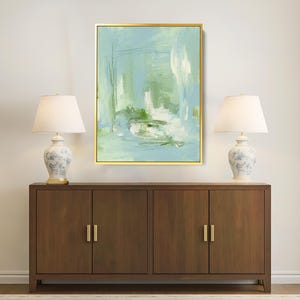 May include: An abstract painting in a gold frame, featuring shades of blue, green, and white. The artwork hangs above a dark wood cabinet with gold handles, flanked by two matching lamps with white shades and floral bases. The lamps provide soft lighting.