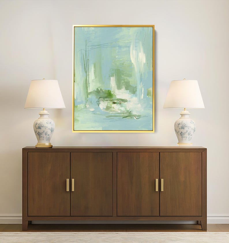 May include: An abstract painting in a gold frame, featuring shades of blue, green, and white. The artwork hangs above a dark wood cabinet with gold handles, flanked by two matching lamps with white shades and floral bases. The lamps provide soft lighting.