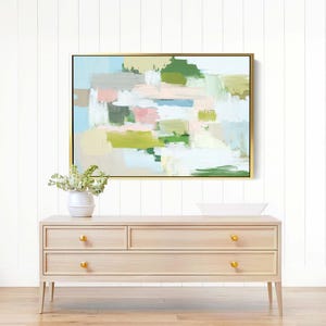 May include: Abstract art painting in a gold frame with pastel colours of green, pink, and blue. The painting hangs above a light wood dresser with gold knobs. A white vase with flowers sits on the dresser.