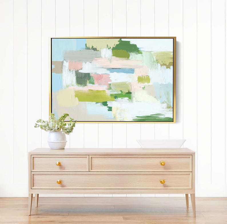 May include: Abstract art painting in a gold frame with pastel colours of green, pink, and blue. The painting hangs above a light wood dresser with gold knobs. A white vase with flowers sits on the dresser.