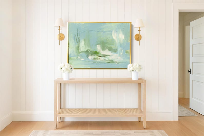 May include: A light wood console table with a woven bottom shelf, topped with white flowers in vases. Above the table hangs a framed abstract painting in shades of blue and green. Gold sconces flank the artwork on a white paneled wall.