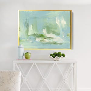 May include: Abstract painting in a gold frame. The artwork features a blend of light blue, green, and white brushstrokes, creating a serene and airy feel. The painting is displayed above a white console table.