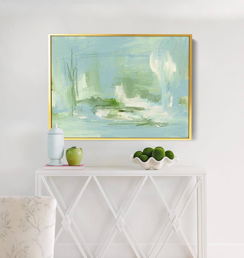 May include: Abstract painting in a gold frame. The artwork features a blend of light blue, green, and white brushstrokes, creating a serene and airy feel. The painting is displayed above a white console table.