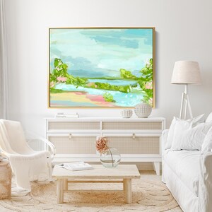 May include: A framed abstract landscape painting with a light blue sky, green trees, and a pink and yellow beach. The painting is hanging on a white wall above a white dresser with a woven rattan top.