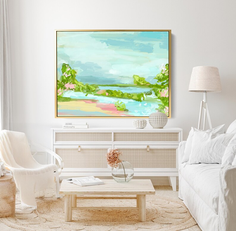 May include: A framed abstract landscape painting with a light blue sky, green trees, and a pink and yellow beach. The painting is hanging on a white wall above a white dresser with a woven rattan top.