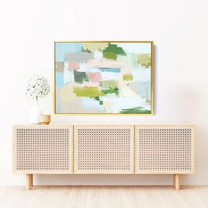 May include: An abstract painting in a gold frame hangs above a light wood cabinet. The painting features pastel colours including pink, green, and blue. A white vase with a flower arrangement and a small wooden bowl sit on top of the cabinet.
