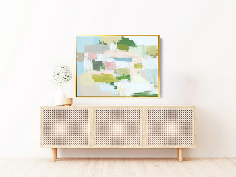 May include: An abstract painting in a gold frame hangs above a light wood cabinet. The painting features pastel colours including pink, green, and blue. A white vase with a flower arrangement and a small wooden bowl sit on top of the cabinet.