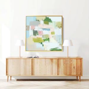 May include: Abstract art print in a gold frame. The artwork features brushstrokes of green, pink, blue, and beige. The painting hangs above a light wood cabinet with two lamps and a book.