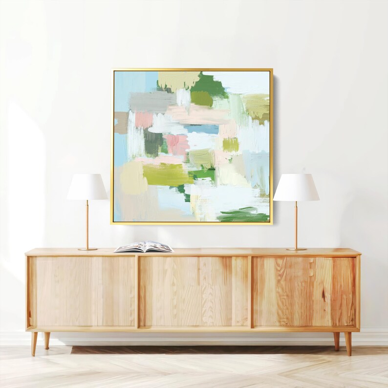 May include: Abstract art print in a gold frame. The artwork features brushstrokes of green, pink, blue, and beige. The painting hangs above a light wood cabinet with two lamps and a book.