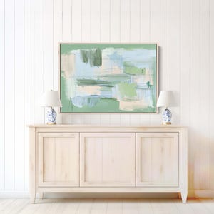 May include: Abstract painting in a wooden frame with green, blue, and beige brushstrokes. It hangs above a light-coloured wooden cabinet, with two white lamps with blue and white patterned bases on either side.