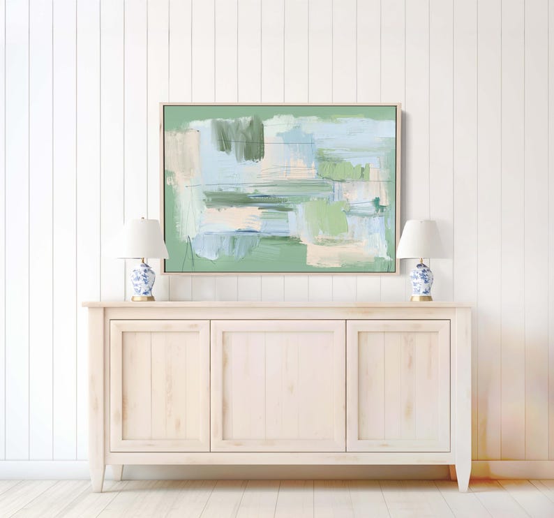 May include: Abstract painting in a wooden frame with green, blue, and beige brushstrokes. It hangs above a light-coloured wooden cabinet, with two white lamps with blue and white patterned bases on either side.