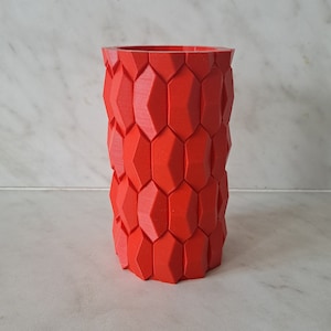 May include: A red, 3D printed cylindrical vase with a geometric pattern. The vase is made of a textured material and has a hexagonal design.