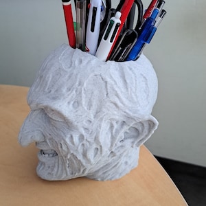 Peut inclure: Un organisateur de bureau en céramique blanche en forme de tête humaine avec une surface texturée. L'organisateur est rempli de stylos et de crayons, y compris du rouge, du bleu et du noir.