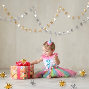 May include: A baby girl wearing a pink and blue tutu and a party hat sits on a white floor with a wrapped gift in front of her.  Silver and gold star garlands hang from the ceiling.