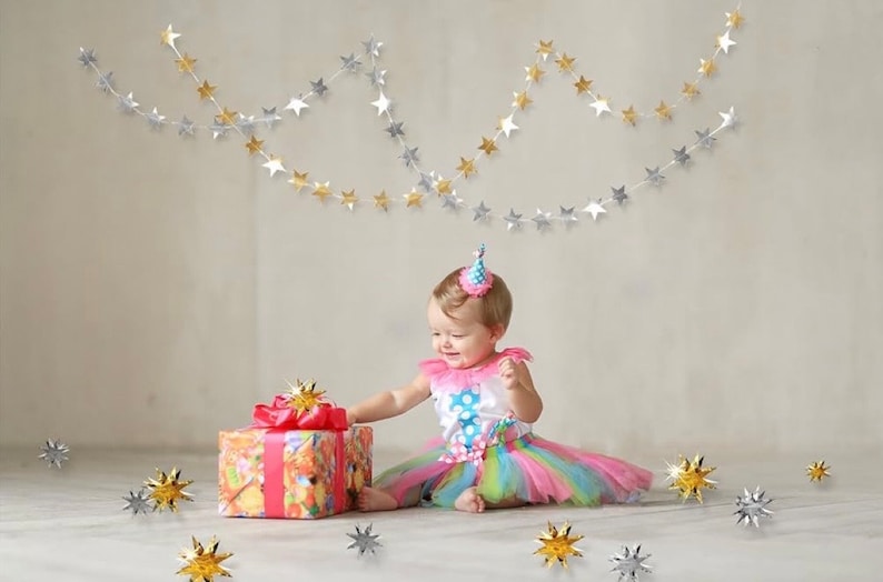May include: A baby girl wearing a pink and blue tutu and a party hat sits on a white floor with a wrapped gift in front of her.  Silver and gold star garlands hang from the ceiling.