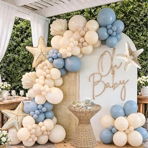 May include: A balloon arch featuring cream and dusty blue balloons, two large starfish-shaped foil balloons, and a white arched backdrop with the wooden text Oh Baby. A wicker pedestal table stands in front with decorative seashells on top.