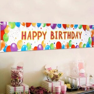 Puede incluir: Una pancarta blanca con globos de colores y el texto "Happy Birthday" en letras rojas y amarillas. La pancarta está colgada en una pared blanca.