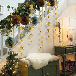 May include: Gold star garland decorations hanging on a white staircase with a white fur throw on a bench. The garland is made of paper stars and is strung on a white string. There are also some paper honeycomb balls in the background.