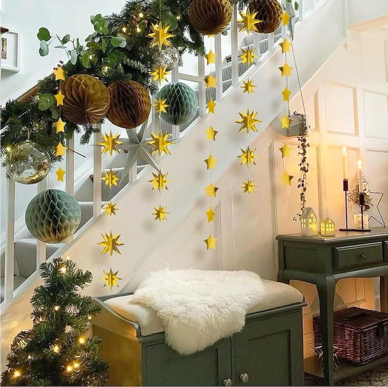 May include: Gold star garland decorations hanging on a white staircase with a white fur throw on a bench. The garland is made of paper stars and is strung on a white string. There are also some paper honeycomb balls in the background.