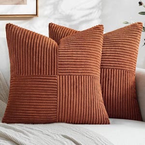 May include: Two square throw pillows in a rich rust-orange color. Each pillow features a textured corduroy fabric with a distinct pattern of alternating vertical and horizontal stripes arranged in a grid-like design across the front surface.