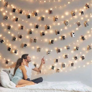 May include: A couple sits on a white bed, looking up at a wall decorated with string lights and photos. The lights are warm white and the photos are hung with clothespins.