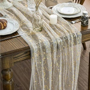 May include: A sheer white table runner with gold foil accents, draped over a wooden table. The runner features vertical stripes and is decorated with a candle, plates, and a small glass vase. A salt shaker and silverware are also visible.