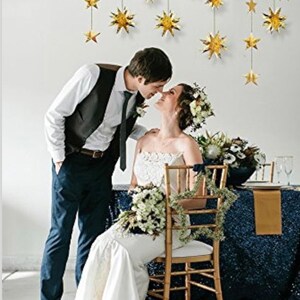 May include: A bride and groom are kissing in front of a white wall decorated with gold star cutouts. The bride is wearing a white wedding dress and the groom is wearing a black suit with a white shirt and a black tie. The bride is holding a bouquet of white flowers. The couple is standing in front of a table with a blue sequined tablecloth.