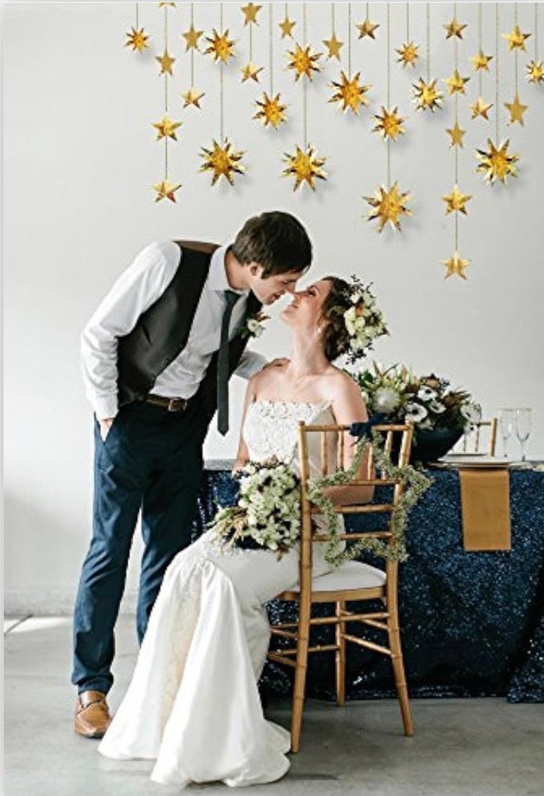 May include: A bride and groom are kissing in front of a white wall decorated with gold star cutouts. The bride is wearing a white wedding dress and the groom is wearing a black suit with a white shirt and a black tie. The bride is holding a bouquet of white flowers. The couple is standing in front of a table with a blue sequined tablecloth.