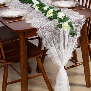 Puede incluir: Camino de mesa de encaje blanco con borde festoneado, adornado con rosas blancas y hojas verdes. El camino de mesa está colocado sobre una mesa de madera oscura con platos y cubiertos dorados. Las sillas de madera rodean la mesa.