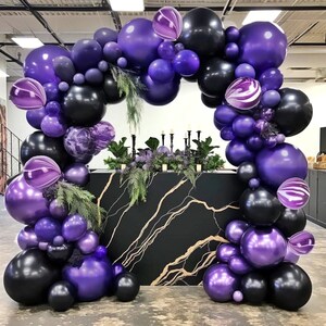 May include: A purple and black balloon arch with greenery, perfect for a party or event decoration. The arch is made of a variety of balloons, including some with a marbled pattern.