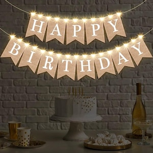 May include: A brown burlap banner with white letters that reads "Happy Birthday" with white lights strung between the letters. The banner is hanging on a brick wall.