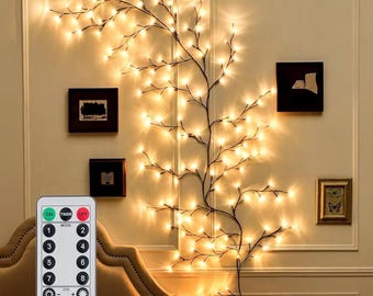 Op de afbeelding: Een wandlamp in de vorm van een tak met talloze kleine, warmwitte ledlampjes bevestigd aan donkere, slanke stelen. De lamp wordt geleverd met een rechthoekige afstandsbediening met knoppen voor aan, uit, timer, acht standen en dimniveaus.