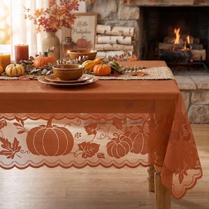 May include: A rectangular wooden dining table covered with a sheer orange lace tablecloth featuring a repeating pattern of pumpkins and autumn leaves. The table is set with brown ceramic bowls, small pumpkins, and a burlap runner.