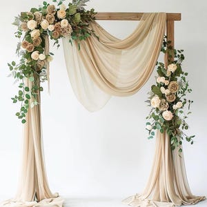 May include: A wooden wedding arch draped with flowing beige fabric and adorned with floral arrangements. The flowers are a mix of cream, peach, and brown roses, with green foliage accents. The arch is set against a white backdrop, creating an elegant display.