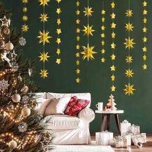 May include: Gold star garland decorations hanging on a green wall with a Christmas tree, a white couch, and a side table with a cup and a small Christmas tree on top.