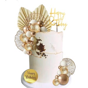 Könnte beinhalten: Ein weißer Kuchen, der mit goldenen und weißen Luftballons, Blattgold und einem goldenen "Happy Birthday"-Topper verziert ist. Der Kuchen steht auf einem weißen Kuchenständer.