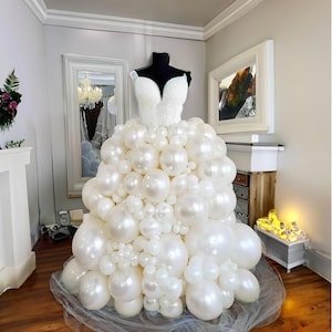 May include: A white wedding dress made entirely of white balloons. The dress is on a mannequin and is displayed in a room with white walls and a fireplace.