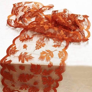 Puede incluir: Un camino de mesa naranja transparente con un patrón repetitivo de hojas de otoño detalladas. La tela tiene una textura de malla delicada y está terminada con un borde festoneado que sigue el contorno de los motivos de hojas en el diseño.