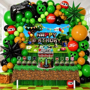 May include: A Minecraft-themed birthday party decoration set featuring a balloon arch in green, orange, black, and red, a backdrop with pixelated landscape, a cake topper, and cupcake toppers with game-inspired characters and TNT motifs.
