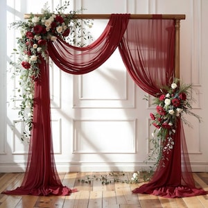 May include: A wooden wedding arch draped with flowing burgundy fabric and adorned with floral arrangements of red and white roses, greenery, and trailing vines. The arch is set against a white wall.