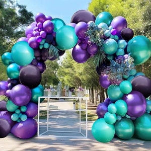 May include: A balloon arch made of purple, teal, and black balloons. The arch is decorated with greenery and is set up in front of a white table.